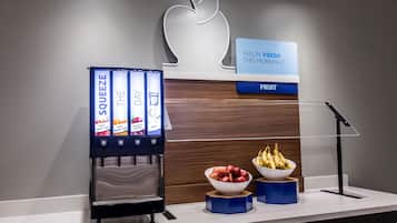 Dispensador de zumos con opciones de manzana, naranja, arándano y agua. Hay cuencos con manzanas y plátanos en la encimera.