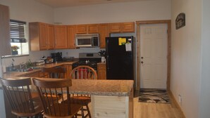 A kitchen with wooden cabinets, a black refrigerator, a microwave, and a stove. There are two wooden chairs and a small island with a countertop.