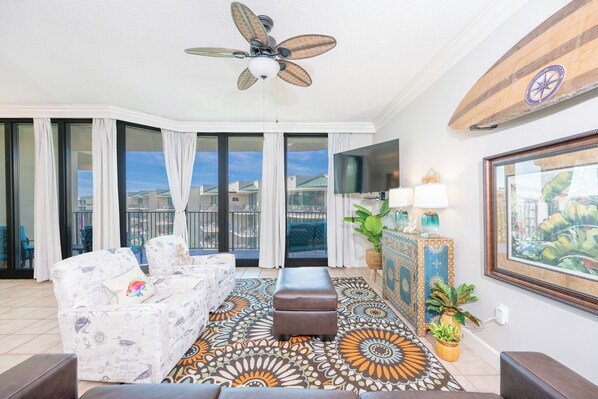 A spacious living room with a ceiling fan, a patterned rug, a sofa, a coffee table, and a large window offering a view of the city. - Phoenix On The Bay 1521 Bay View (Orange Beach)