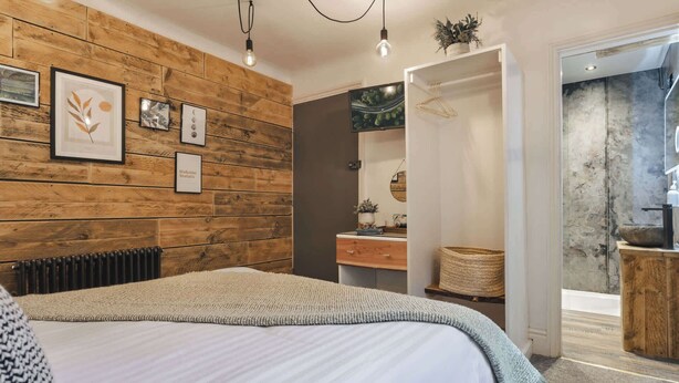 Quarto com parede de madeira, cama com edredom branco, mesa de cabeceira com abajur e luminária pendente.