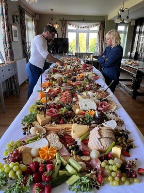 Een lange tafel vol met verschillende kazen, vleeswaren, fruit en groenten in een goed verlichte kamer met grote ramen.