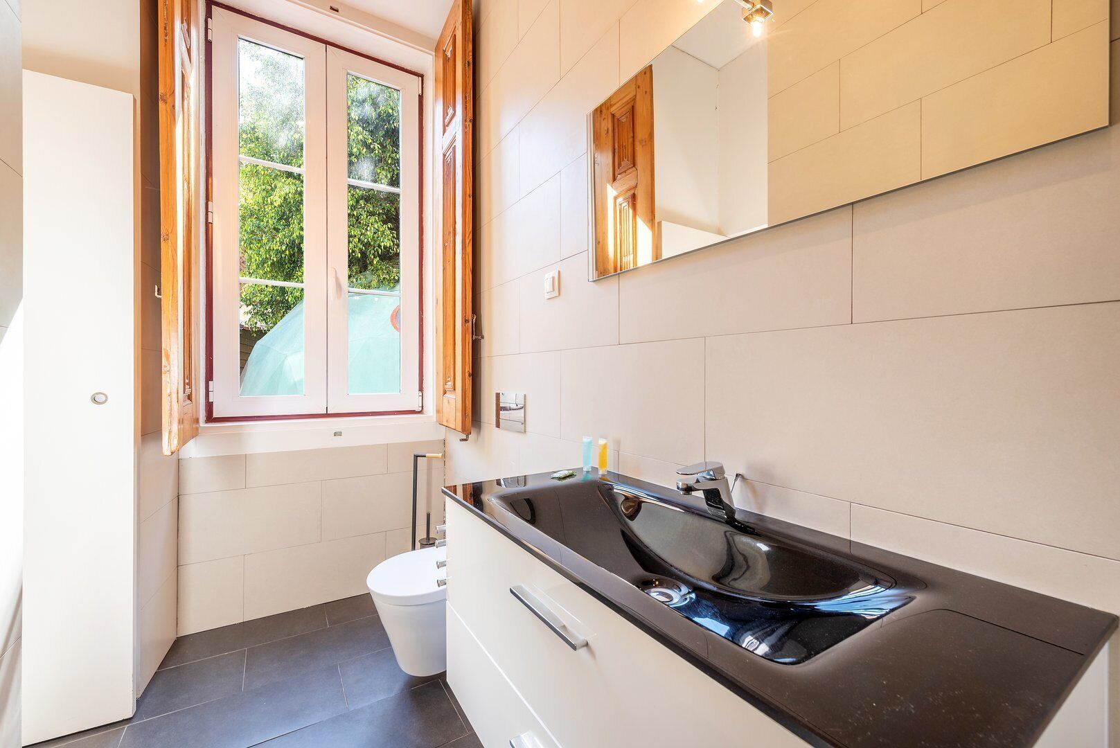Un baño moderno con un lavamanos grande, un inodoro y una ventana con contraventanas de madera.
