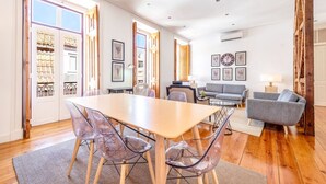 Ein geräumiges Wohnzimmer mit einem Holzesstisch, klaren Plastikstühlen, einem grauen Sofa und einem großen Fenster, das für natürliches Licht sorgt.