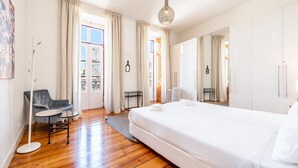 Ein modernes Hotelzimmer mit einem großen Bett, einer Sitzecke mit Stuhl und Tisch, einem Balkon mit Vorhängen und einer hängenden Leuchte.