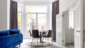 Een kamer met een blauwe bank, een ronde tafel met stoelen en uitzicht op gebouwen door een groot raam.