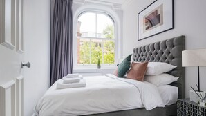 Een net opgemaakt bed met witte lakens en een grijs hoofdeinde, een raam met gordijnen en een lijst met een afbeelding aan de muur.