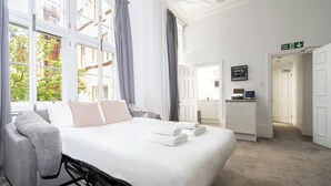 Habitación de hotel con una cama grande, un sofá y un escritorio. Se aprecia una ventana con cortinas, una puerta con un letrero y una señalización de salida de incendios en la pared.