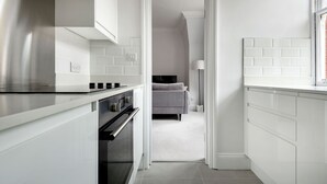 A modern kitchen with white cabinetry, a built-in oven, and a stainless steel range hood. A hallway leads to a bedroom with a grey sofa and a bedside table. - The Cadogan Suite next to Sloane Square (London)