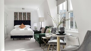 Een moderne hotelkamer met een bed, een groene fauteuil, een ronde tafel met een vaas, en uitzicht op de stad door grote ramen.