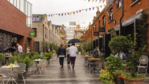 Een voetgangersstraat met terrasjes en plantenbakken, waar mensen lopen en dineren.