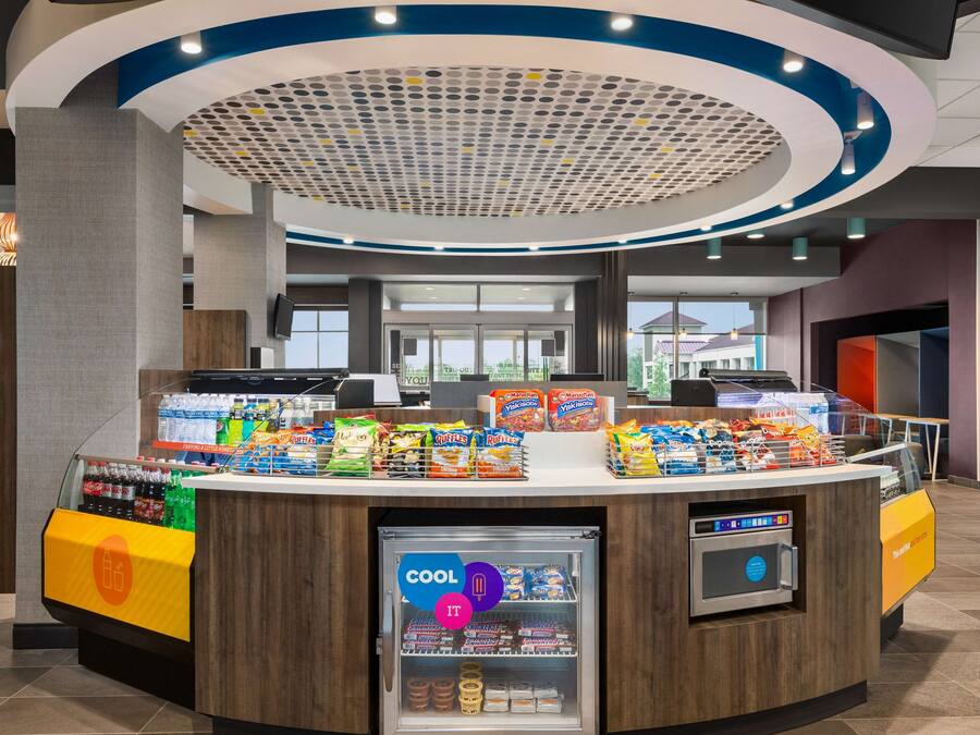 Un área de autoservicio con bebidas y aperitivos variados, una sección refrigerada y un terminal de pago digital.