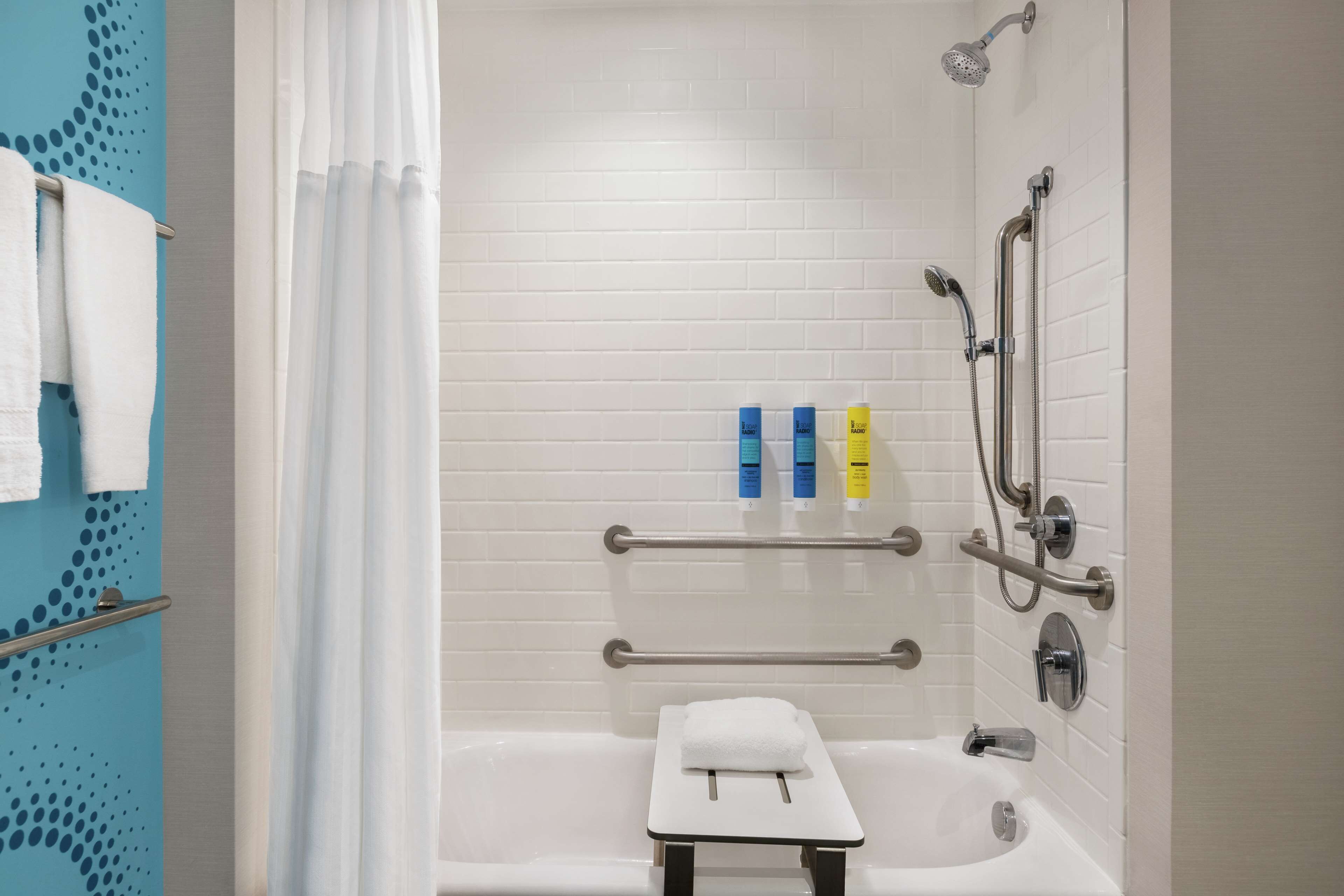 A modern bathroom with a white bathtub, a small stool, a shower with a handheld showerhead, and a handrail.