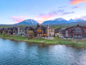 Un albergue junto al lago con varias construcciones, una chimenea de piedra y vistas a montañas nevadas al fondo.