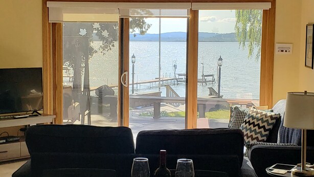 Ein Wohnzimmer mit großem Fenster mit Seeblick, einer Couch, einem Tisch mit Weingläsern und einem Fernseher.