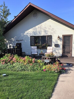 Une maison avec un toit en pente, un porche à l’avant avec deux chaises, une petite table et un parterre fleuri avec différentes plantes et fleurs.