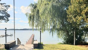 Ein Uferweg an einem See mit einem hölzernen Steg, der ins Wasser ragt, umgeben von Bäumen und Laternen.