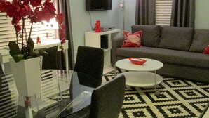 A modern living room with a glass dining table, a grey sofa, a flat-screen TV, and a patterned rug. - Beautiful Studio on Collins Ave by the Beach with Private Parking (Miami Beach)