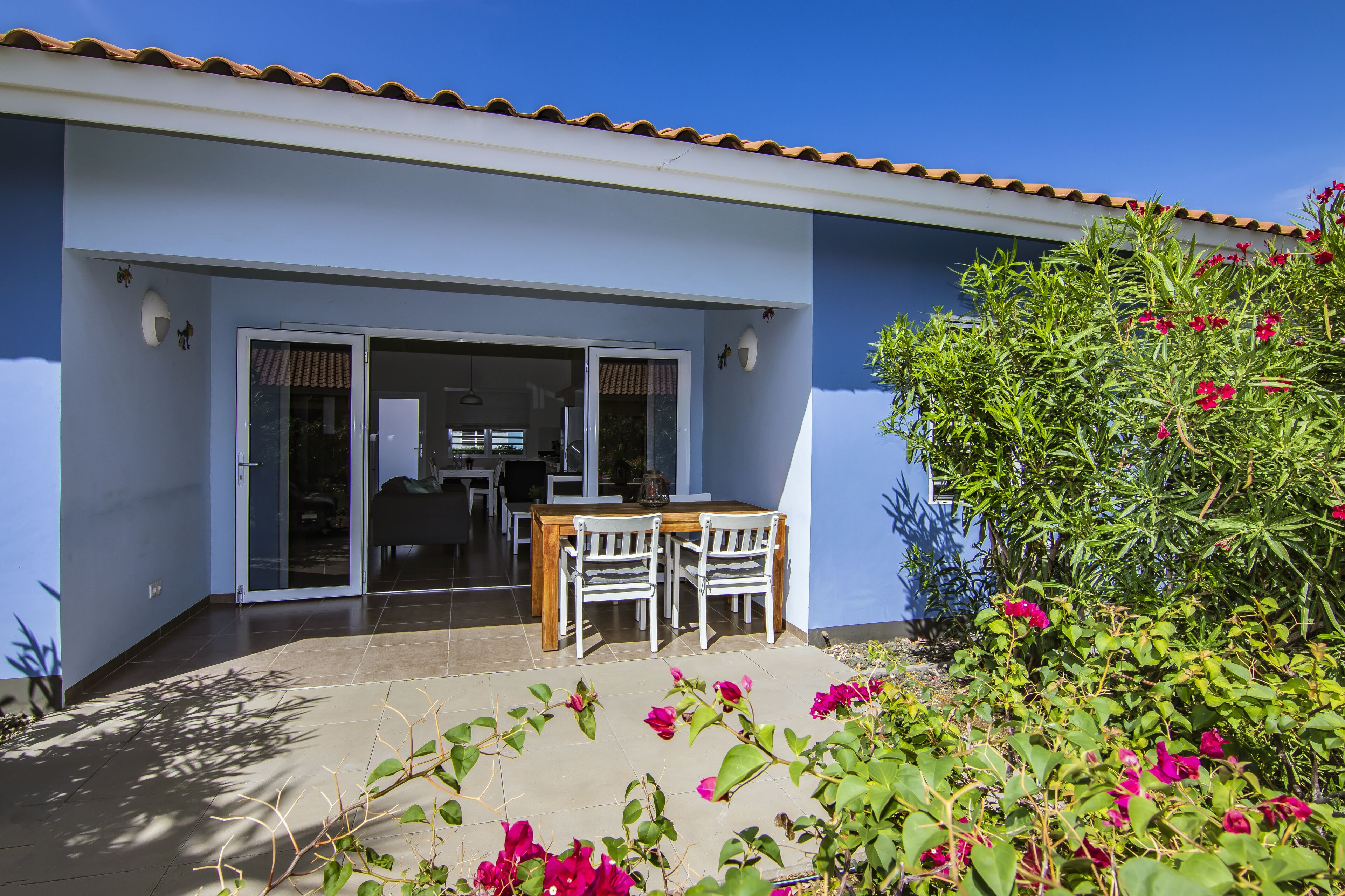 Une maison moderne dotée d’un patio, d’un coin repas avec une table et des chaises, et d’un jardin agrémenté de fleurs roses.