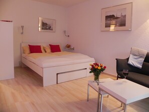 A bedroom with a bed, a bedside table with a vase of flowers, a framed picture on the wall, and a sofa with a glass of wine. - "Garden I" Studio, underground parking, balcony, central location, clinic within walking distance (EK/OSK) (Ravensburg)