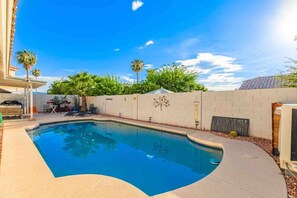 Ein Pool mit geschwungener Kante, umgeben von einer niedrigen Mauer und einer Terrasse mit Gartenmöbeln.