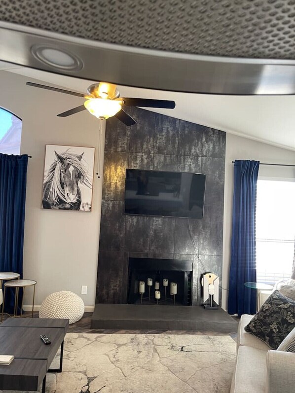 Ein modernes Wohnzimmer mit einem Flachbildfernseher an einer dunklen Wand, einem Deckenventilator, einem Kamin mit Kerzen und einem großen Fenster mit Vorhängen.