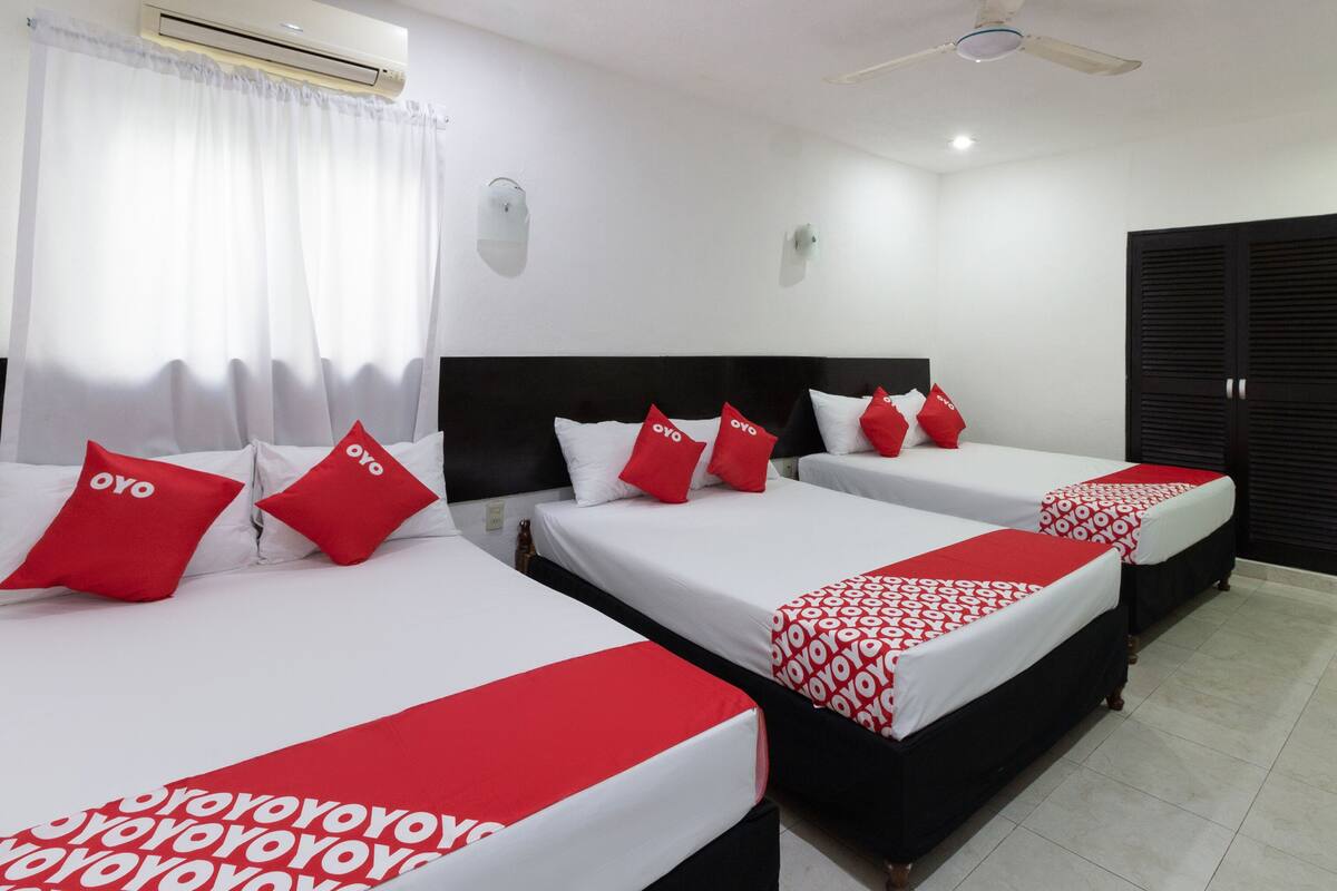 A hotel room with three beds, each with red and white bedding and pillows, a ceiling fan, and an air conditioning unit.