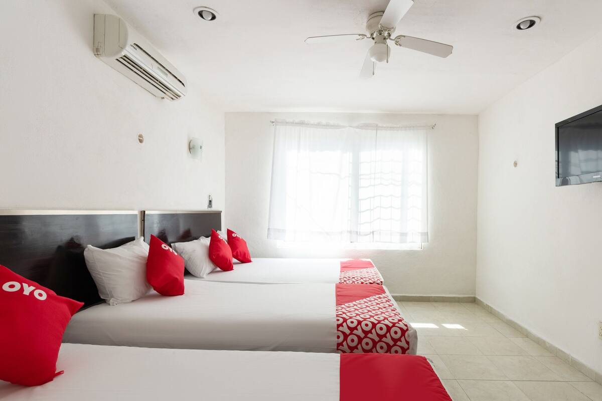 A hotel room with two beds, a ceiling fan, an air conditioning unit, a window with sheer curtains, and a flat-screen TV mounted on the wall.