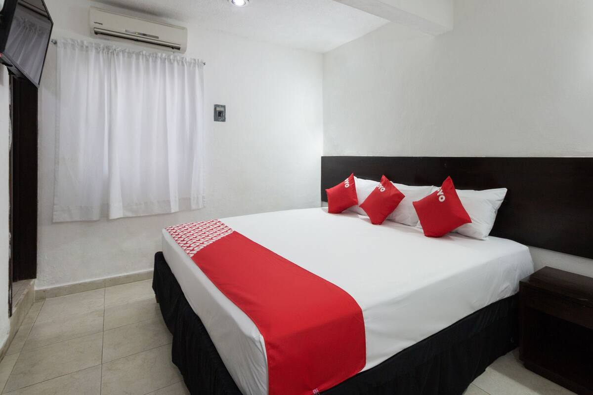 A hotel room with a bed, a nightstand, a TV, and an air conditioning unit.