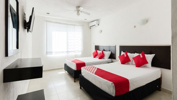 Habitación de hotel con dos camas, un escritorio, un espejo, un ventilador de techo y una ventana con cortinas traslúcidas.