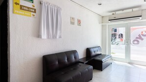 Zona de recepción de un hotel con un área de espera que cuenta con dos sofás de cuero negro y una cortina blanca. Hay señales indicativas de la ruta de evacuación y del primer piso.