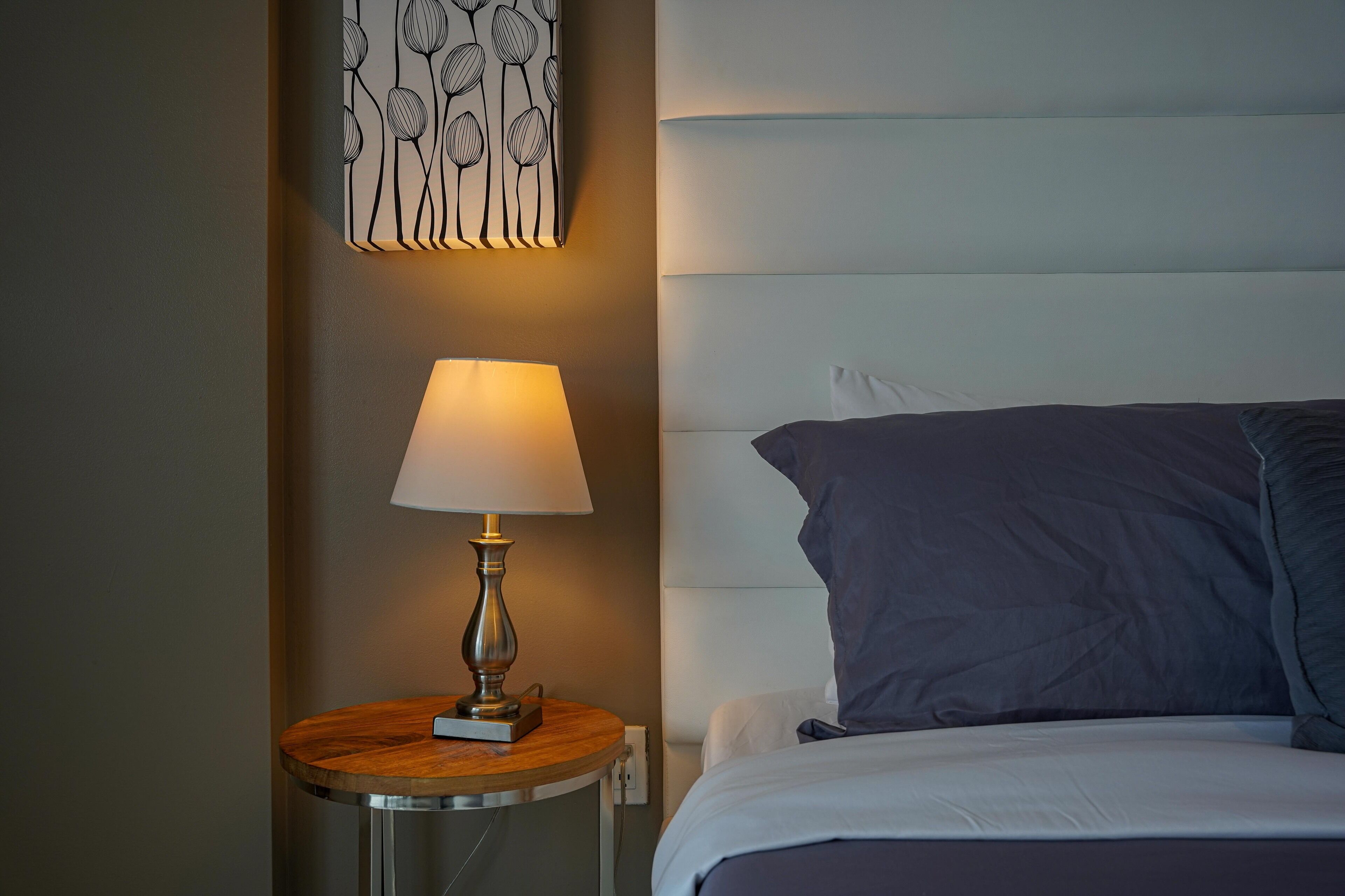 A neatly made bed with a dark blue pillow and a white headboard. A round wooden nightstand with a lamp is positioned to the left of the bed.