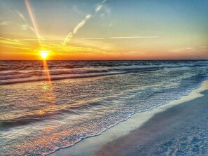 A beach with the sun setting over the ocean, creating a reflection on the water.