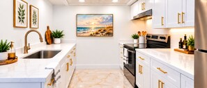 Une cuisine moderne avec des meubles blancs, des poignées dorées, un évier en acier inoxydable et un tableau représentant une scène de plage.