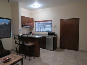 Una cocina compacta con refrigerador, microondas y ventana con persianas. Hay dos taburetes de bar y una mesa de comedor con un tazón.