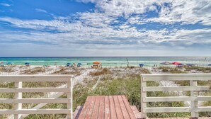 Blick auf einen Strand mit einer Holzterrasse, Strandsonnenschirmen und Menschen, die das Meer genießen.