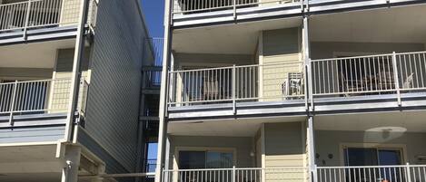 A multi-story residential building with balconies, a parking area in front, and a clear sky.