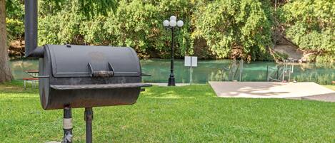 Una parrilla para barbacoa sobre una base de concreto en una zona con césped, con un estanque y árboles al fondo.