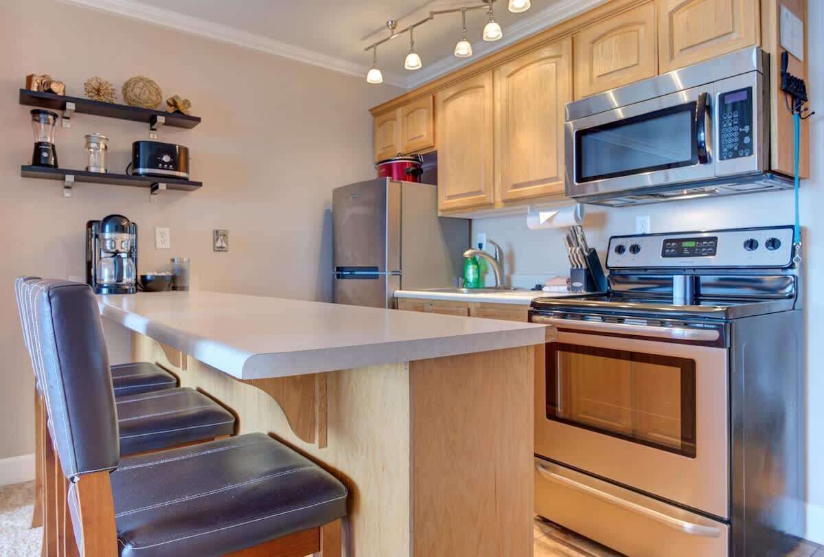 Een moderne keuken met een centraal kookeiland, roestvrijstalen apparatuur, houten kastjes en een barkruk.
