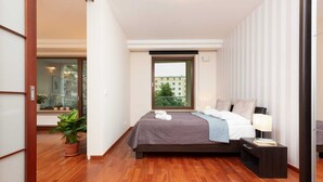 A modern bedroom with a large bed, a nightstand, a potted plant, and a window with a city view. - Modern Apartment in the Center | Balcony | Parking (Warszawa)
