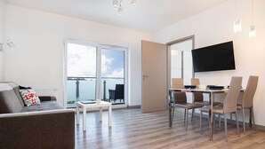 A modern living room with a sofa, dining table, and chairs, a flat-screen TV mounted on the wall, and a sliding glass door leading to a balcony.