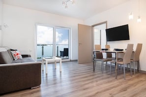 A modern living room with a sofa, dining table, and chairs, a flat-screen TV mounted on the wall, and a sliding glass door leading to a balcony.