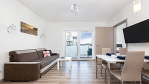 A modern living room with a sofa, a coffee table, a dining table with chairs, a flat-screen TV mounted on the wall, and a large window with a view of buildings.