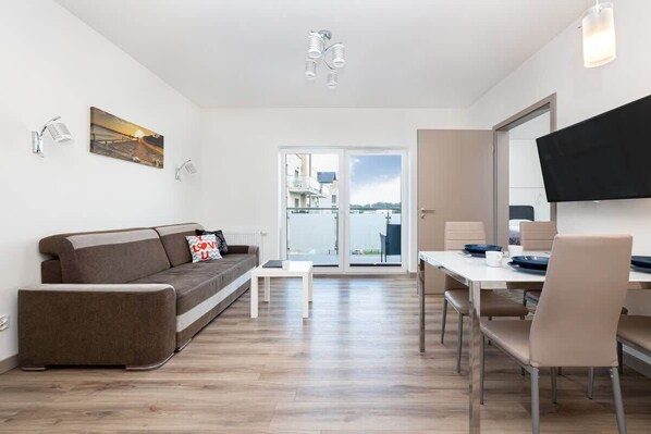 A modern living room with a sofa, a coffee table, a dining table with chairs, a flat-screen TV mounted on the wall, and a large window with a view of buildings.