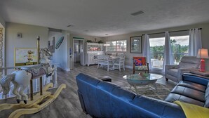 Amplia sala de estar con un sofá azul, una mesa de centro de vidrio y un caballito de madera. Hay un comedor con muebles blancos y una cocina al fondo.