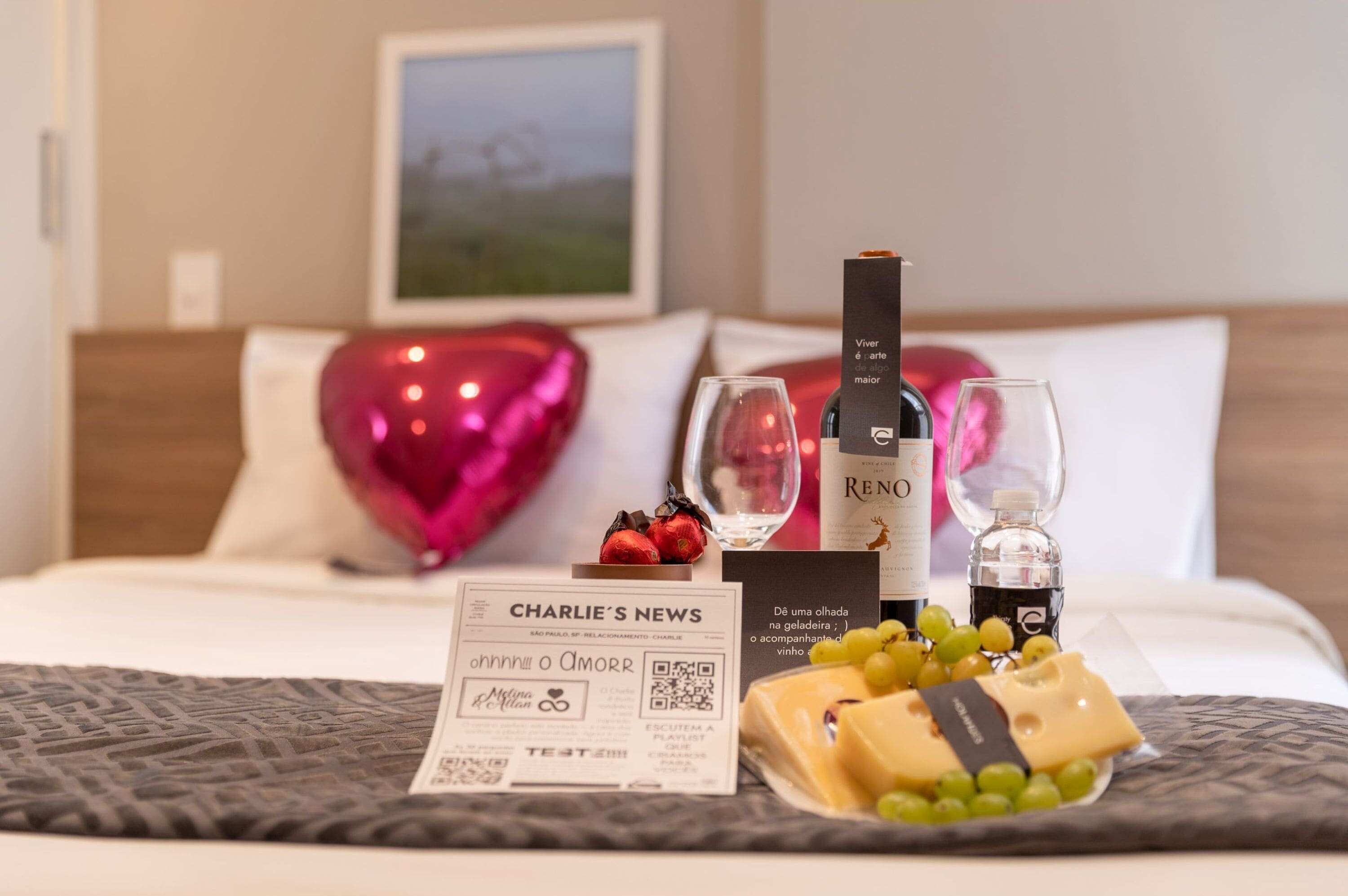 Une chambre d’hôtel bien rangée, avec un lit, une décoration en forme de cœur, une bouteille de vin, des verres, un menu et une assiette de fromage et de raisins.