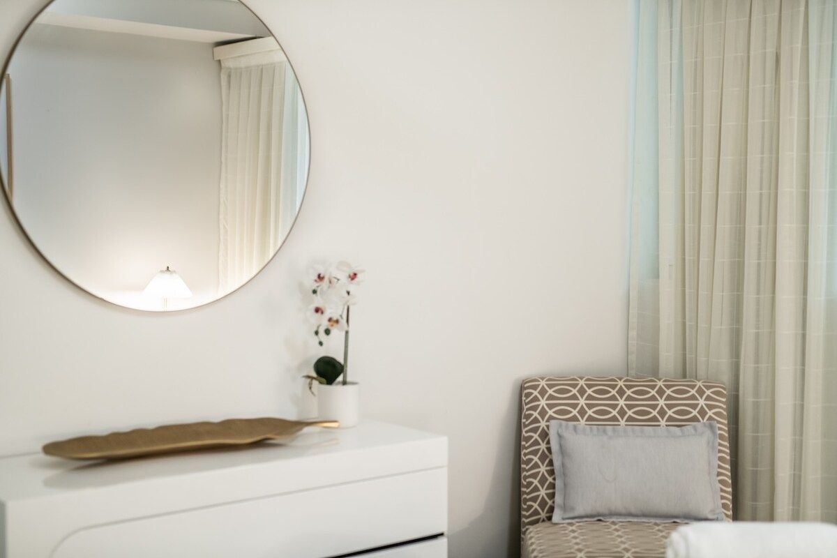 A bedroom with a bed, a bedside table, a mirror, and a window with curtains.