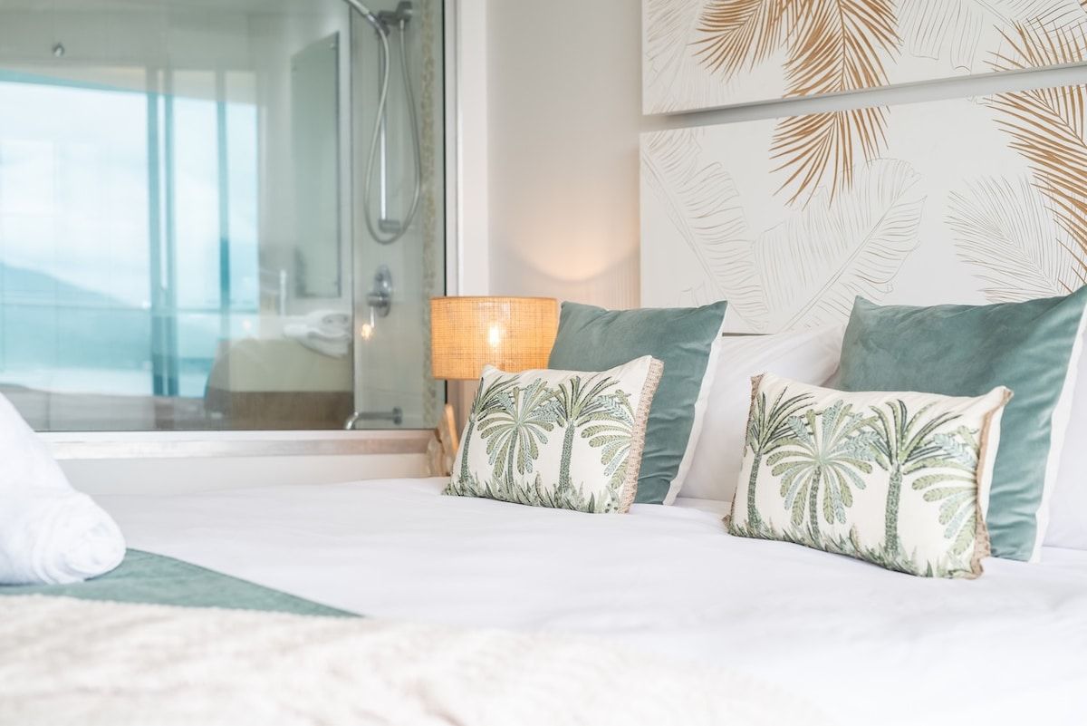 Une chambre d’hôtel avec un lit, deux oreillers ornés de motifs de palmiers, une lampe de chevet et une vue sur un balcon.