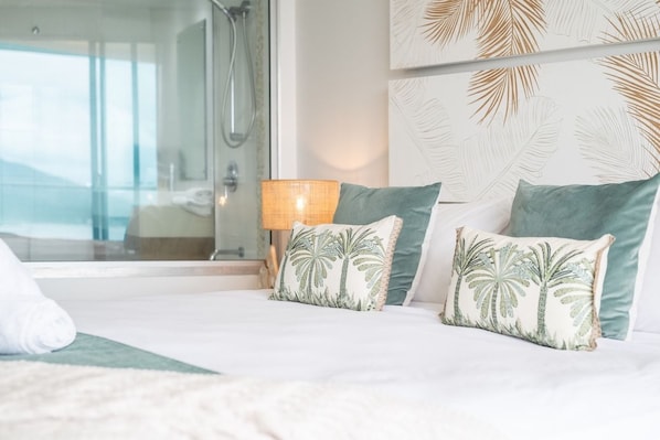 Una habitación de hotel con una cama, dos almohadas con diseño de palmeras, una lámpara de noche y vistas a un balcón.