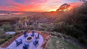 Uma área de estar circular ao ar livre, com cadeiras e lareira, cercada por vegetação nativa e vista para o pôr do sol.