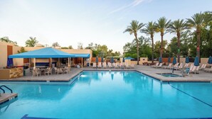 A swimming pool with lounge chairs and umbrellas, surrounded by palm trees and a covered seating area.